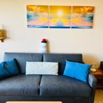 Ein grauer Sofa mit blauen und weißen Kissen. Wandbild zeigt Sonnenuntergang mit Segelboot.