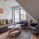 Wohnzimmer mit Sofa, zwei Sesseln, Tisch und Fenster. Wandbild und Pflanzen sind sichtbar.