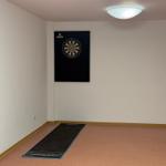 Dartwandschutz an der Wand, schwarzes Dartboard, dunkler Bodenbelag mit blauer Kante.