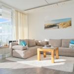 Wohnzimmer mit Ecksofa, Holztisch und Wandbild. Große Fenster mit Blick nach draußen.