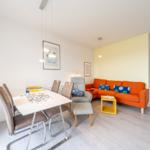 Essbereich mit Tisch und Stühlen. Wohnbereich mit orangefarbenem Sofa und gelbem Beistelltisch.