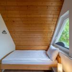 Ein Schlafzimmer mit Holzdecke, Bett und Fenster. Draußen sind Bäume sichtbar.