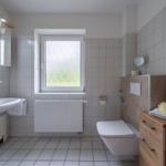 Badezimmer mit Waschbecken, Toilette und Fenster. Holzschrank und Spiegel.