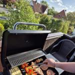 Grill mit Fleisch und Gemüse auf Terrasse mit Sicht auf Grün und Häuser.