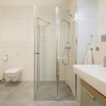 Badezimmer mit Dusche, Waschbecken und Toilette. Holzregal und graue Fliesen.