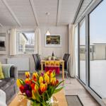 Wohnbereich mit Esstisch, Sofa und großen Fenstern. Blumen und TV sind sichtbar.