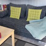 Ein grauer Sofa mit zwei gelben Kissen und einer hellblauen Decke. Holztisch mit Buch und Schale.