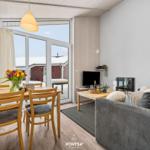Wohn- und Essbereich mit Sofa, Tisch und Stühlen. Durchgang zur Terrasse mit Blick auf rote Gebäude.