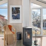 Raum mit Kamin, Sessel und Glasplatte. Große Fenster zeigen Außenbereich.