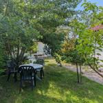 Garten mit Tisch und Stühlen unter Bäumen. Weiße Wand und roter Dachfirst sichtbar.