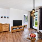 Living room with TV, glass table, and seating. Wooden floor and windows with view.