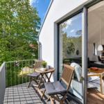 Terrasse mit Tisch und Stühlen, Blick auf grüne Bäume. Durchgehendes Glas führt in Wohnbereich mit Sofa und Tisch.