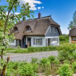 Thatched-roof houses with green gardens and gravel paths under a blue sky.