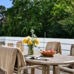 Holztisch mit Stühlen, Blumen und Äpfeln auf Terrasse mit Blick auf Bäume.
