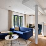 Ein Wohnzimmer mit blauem Sofa, weißem Tisch und Holzboden. Durchgehende Fensterfront mit Blick nach draußen.