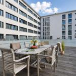 Terrasse mit Holztisch und Stühlen, Blick auf Gebäude mit Glasgeländer.