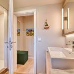 Badezimmer mit Dusche, Waschbecken und Spiegel. Durchgang zu einem Raum mit grünem Sitzkissen.