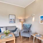 Ein Wohnzimmer mit grauem Sofa, Sessel und Holztisch. An den Wänden hängen Bilder.