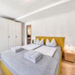 Schlafzimmer mit gelbem Bett, weißen Wänden und Holzboden. Neben dem Bett steht ein Nachttisch.