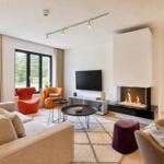 Wohnzimmer mit Sofa, Sesseln, Kamin und Fernseher. Fenster mit Blick auf Grün.