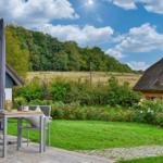 Terrasse mit Tisch und Stühlen, Blick auf grünen Garten und strohgedeckte Häuser.