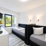 Room with two beds, window with view of green, wall lamp and TV.