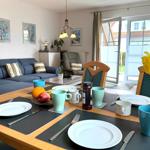 Esstisch mit Tischdecken, Tassen und Obst. Im Hintergrund Wohnzimmer mit Sofa und Glastür.