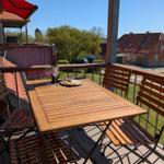 Terrasse mit Holztisch und Stühlen, Blick auf Haus und Wasser.
