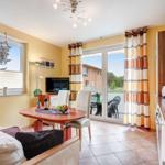Wohn- und Essbereich mit Balkon, Küche und Fernseher. Holztisch, Sofa und Fenster mit Jalousie.
