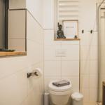 Badezimmer mit Toilette, Duschbereich und Pflanze. Buddha-Statue und Heizkörper sichtbar.