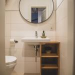Badezimmer mit Waschbecken, Spiegel und Holzregal. Toilette und Duschwand sind sichtbar.