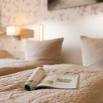 Ein Bett mit Kissen und einem Buch darauf. Wand mit Blumenmuster und Nachttischlampe.