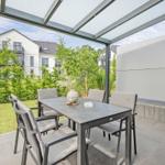 Terrasse mit Tisch und Stühlen unter Glasdach. Grünfläche und moderne Häuser im Hintergrund.
