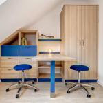 Ein Arbeitszimmer mit Schreibtisch, zwei blauen Stühlen und einem Holzschrank unter Dachschrägen.