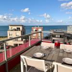 Terrasse mit Tisch, Stühlen und Grill. Blick auf Meer und Gebäude.