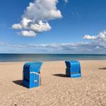 Drei blaue Strandkörbe stehen auf sandigem Strand vor dem Meer unter blauem Himmel mit weißen Wolken.