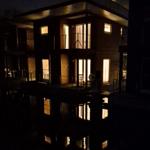 Moderne Holzhäuser mit beleuchteten Fenstern reflektieren sich im Wasser bei Nacht.