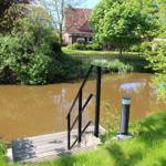 Holzsteg mit Geländer führt zum Ufer des Flusses. Grün umgeben, Haus im Hintergrund.