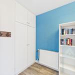Zimmer mit blauer Wand, weißem Schrank und Bücherregal. Holzdielenboden und Heizung.