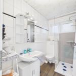 Badezimmer mit Dusche, Waschbecken und Toilette. Weiße Fliesen und Holzboden.