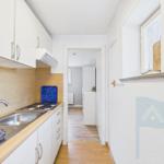 Küche mit Spüle, Kochfeld und Küchenschränken. Blick in den Flur mit Holzboden.
