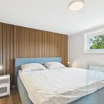 Schlafzimmer mit Bett, Nachttisch und Holzwand. Fenster mit Blick auf Bäume.