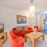 Wohnzimmer mit rotem Sofa, Holztisch und Fernseher. Küche und Schlafzimmer im Hintergrund.