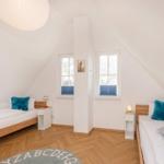 Zwei Betten mit Kissen, zwei Fenster, Holzboden und Deckenlampe in einem Dachzimmer.