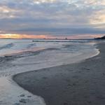 Strand mit Wellen und Himmel bei Sonnenuntergang. Im Hintergrund ist eine Promenade und ein Leuchtturm sichtbar.
