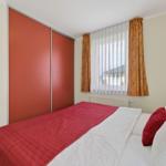 Schlafzimmer mit Bett, rotem Bettbezug, Fenster mit Vorhängen und Schiebetüren.