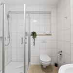 Badezimmer mit Dusche, Toilette und Waschbecken. Weiße Fliesen und Holztür.