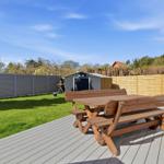 Holztisch und Bänke auf Terrasse mit Garten und Zaun im Hintergrund.