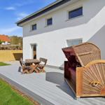 Terrasse mit Holztisch, Bistrostühlen und Strandkorb. Hintergrund: weißes Haus, Holzzaun und Gras.