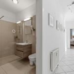 Bathroom with shower, sink, and toilet. Hallway with radiator and view into bedroom.